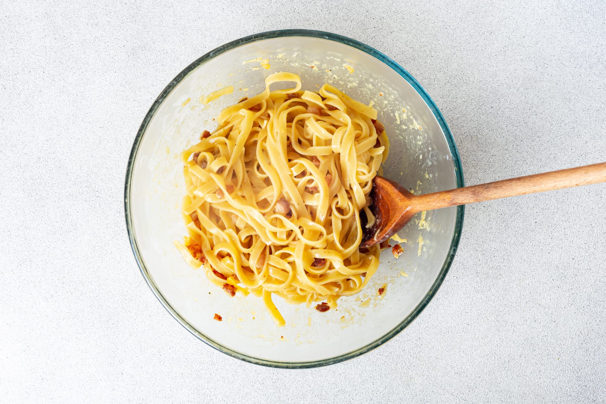 Mixing pasta into pancetta and eggs mix in a glass bowl with a wooden spoon.