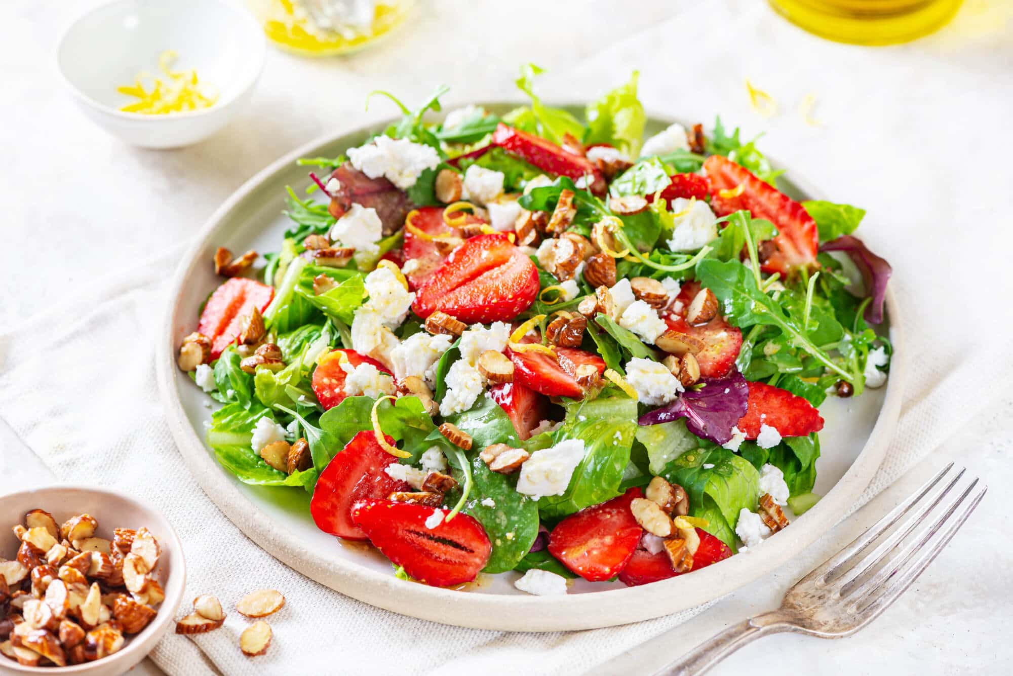 Summer berry salad on a white plate with a fork, salad dressing, and lemon zest on the side.