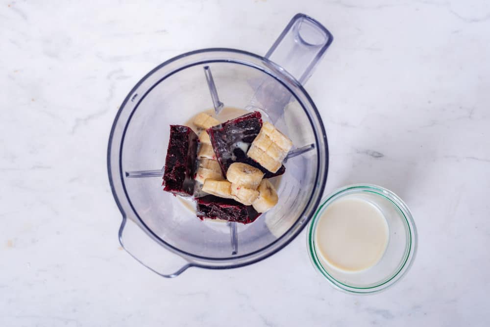 acai smoothie ingredients in a blender bowl with a glass of milk on the side.