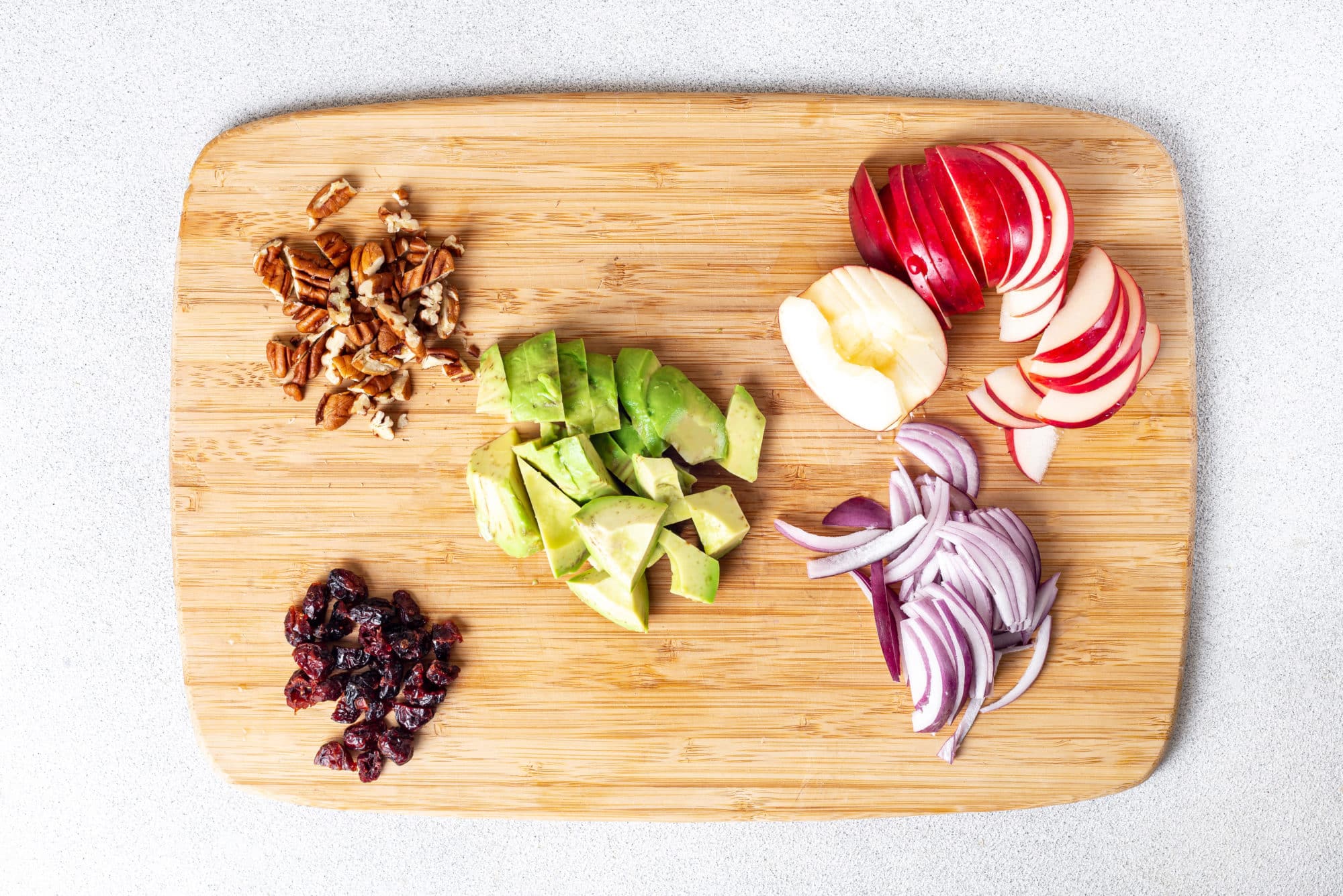 Chopped apple red onion avocado dried cranberries and walnuts on a wooden cutting board.