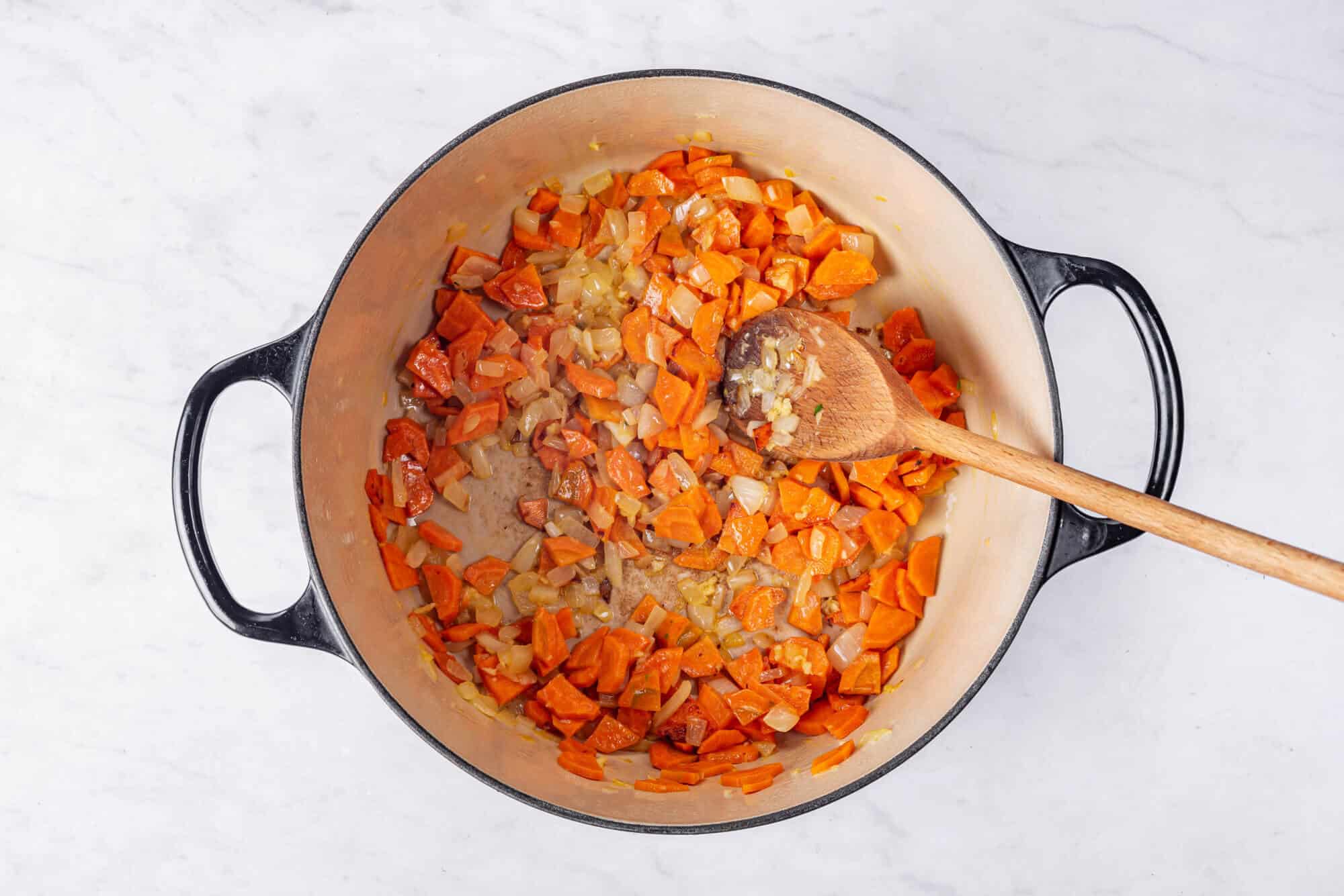 Onions and carrots sauteing in a soup pot with a wooden spoon.