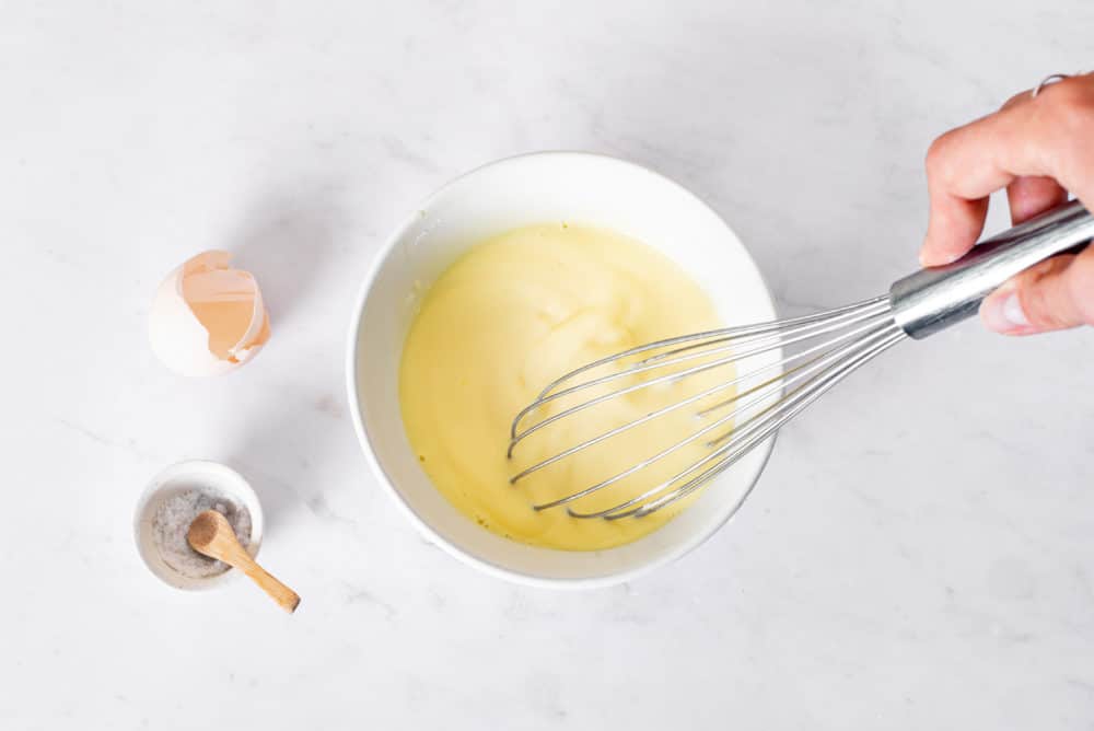 Milk and eggs in a bowl with a whisk and egg shells on the side.