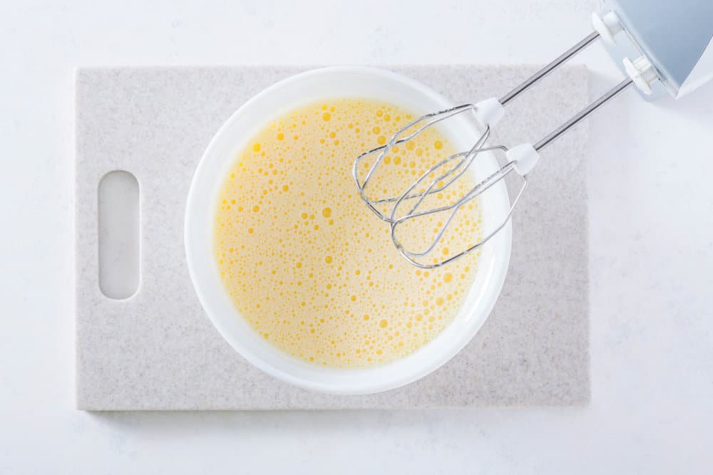 Mandarin mousse cake batter in a white bowl with a cutting board with a hand mixer.