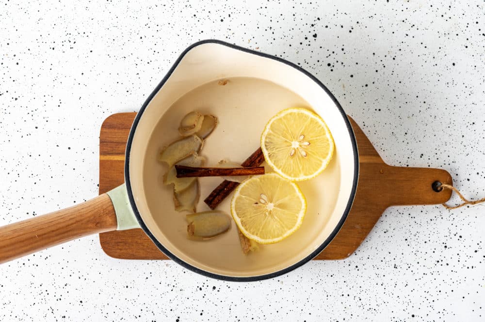 tea brewing in a pot on a wooden board with lemon cinnamon water and ginger slices in the pot.