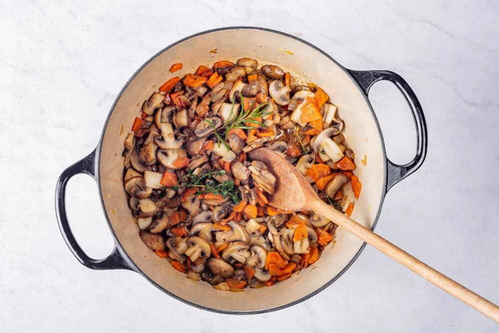 Mushroom soup vegetable ingredients sauteing in a large soup pot with a wooden spoon.