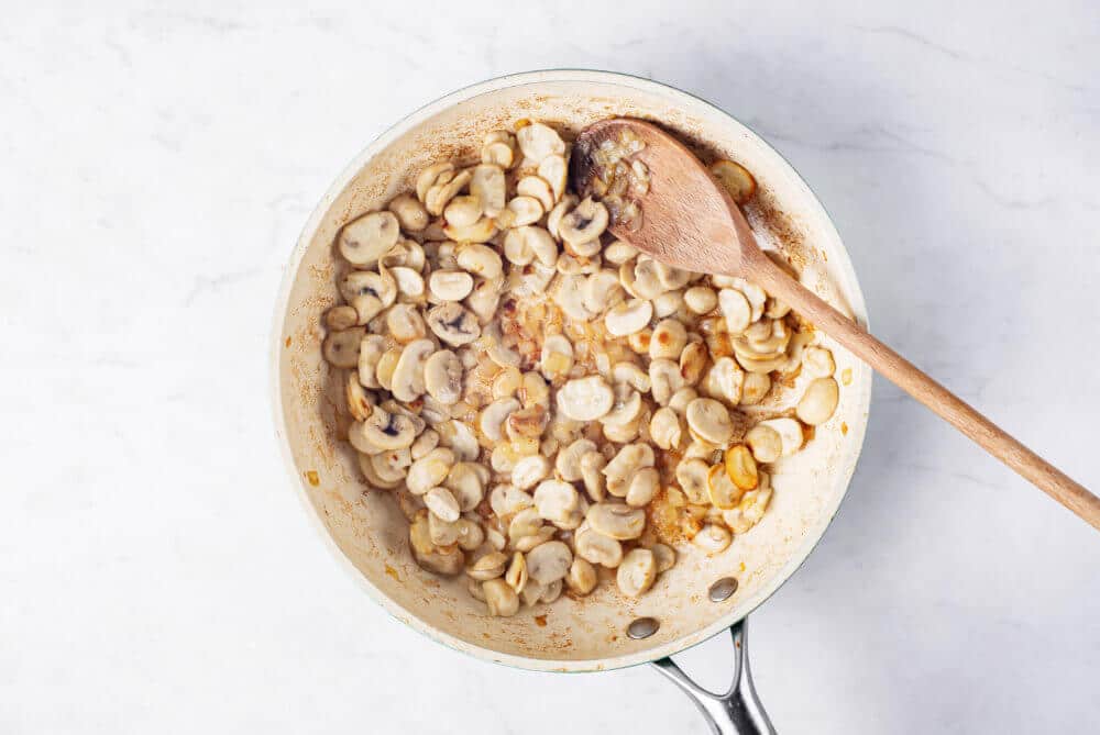 Mushrooms browning in a skillet, mixing with a wooden spoon.