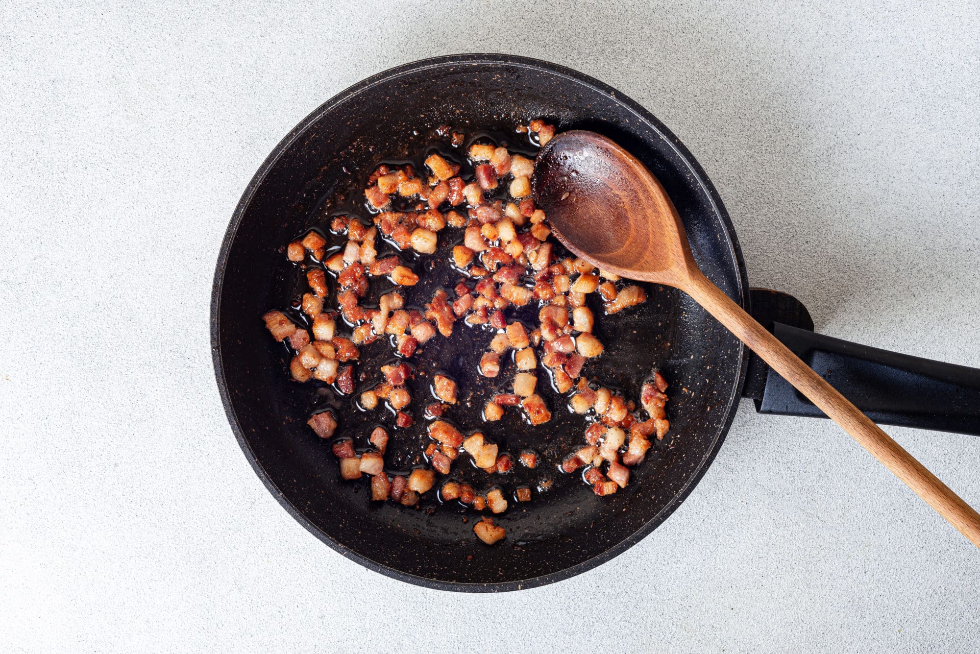 Pancetta sauteing in a skillet with a wooden spoon.