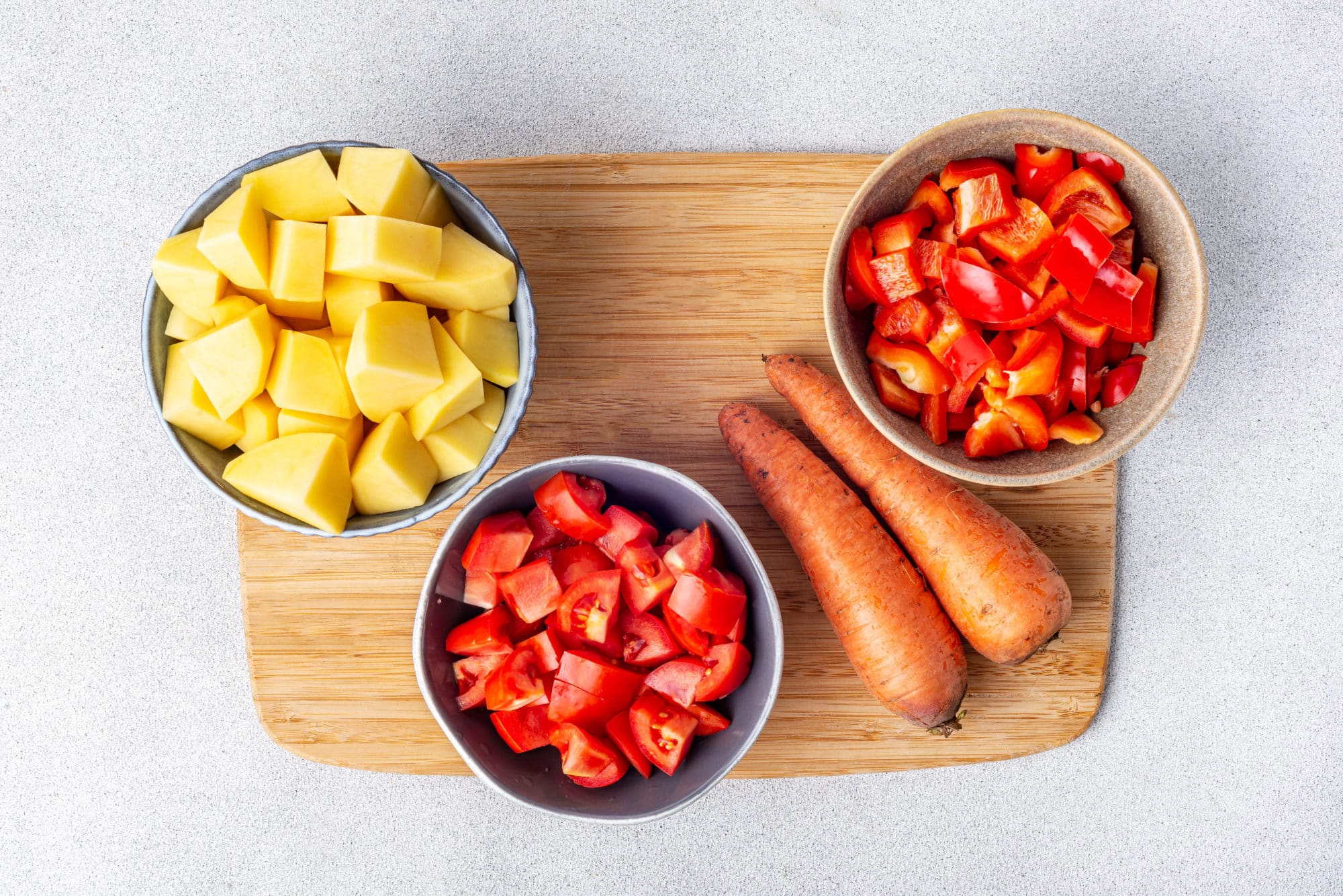 Chopped veggies in bowls on a wooden cutting board chopped potatoes tomatoes and red bell peppers and whole carrots.