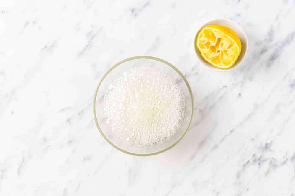 a used lemon wedge on the side of a glass bowl with combined lemon juice and baking soda in it bubbling.