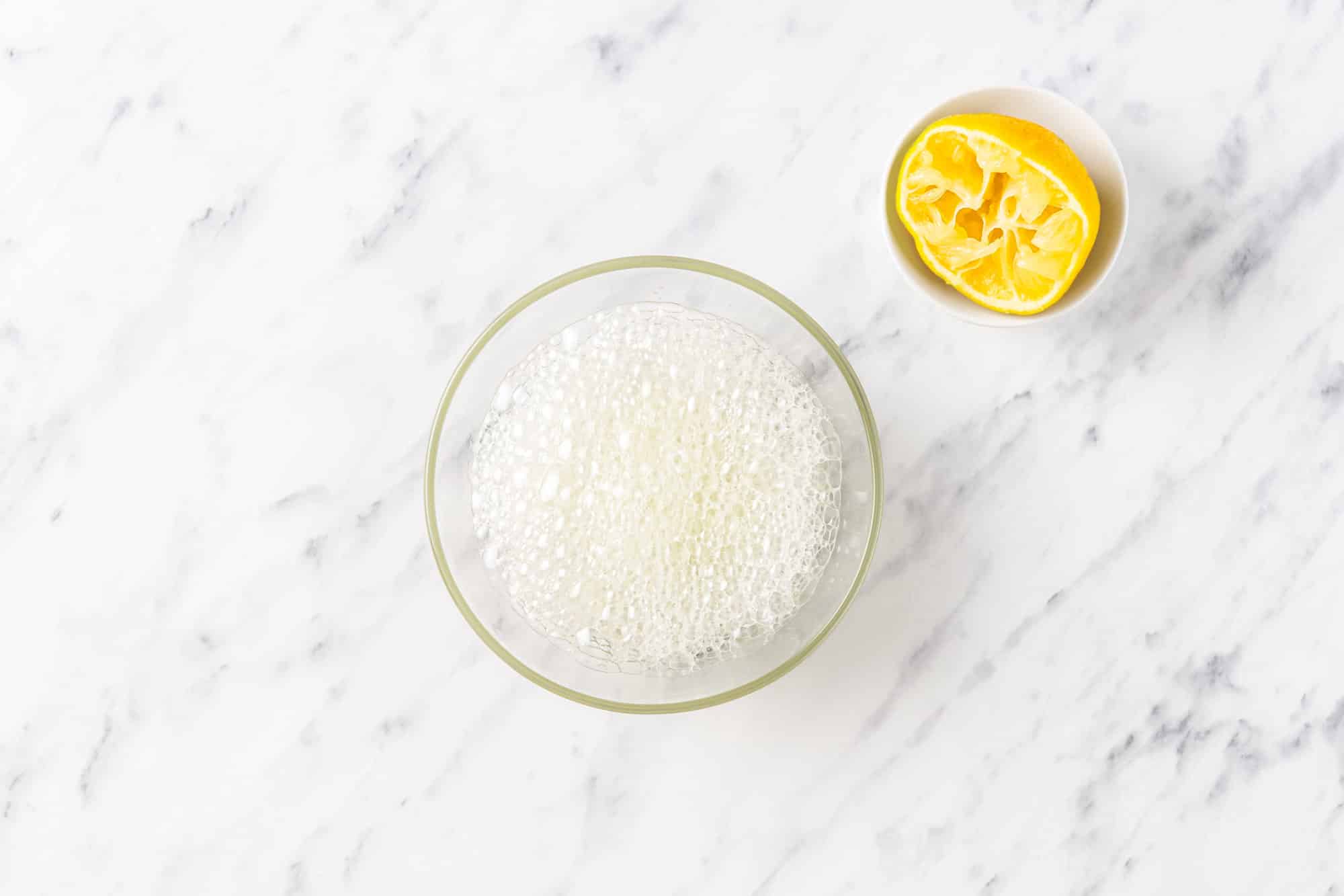 a used lemon wedge on the side of a glass bowl with combined lemon juice and baking soda in it bubbling.