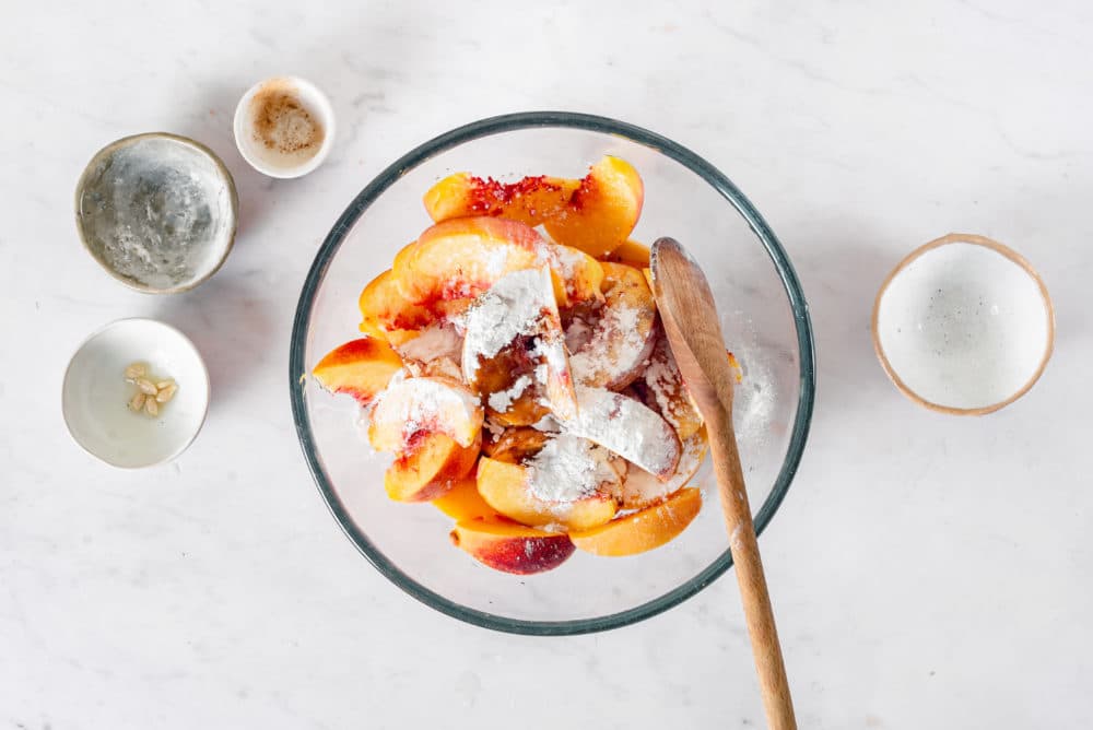 Peaches with flour in a clear bowl with a wooden spoon with bowls on the side.