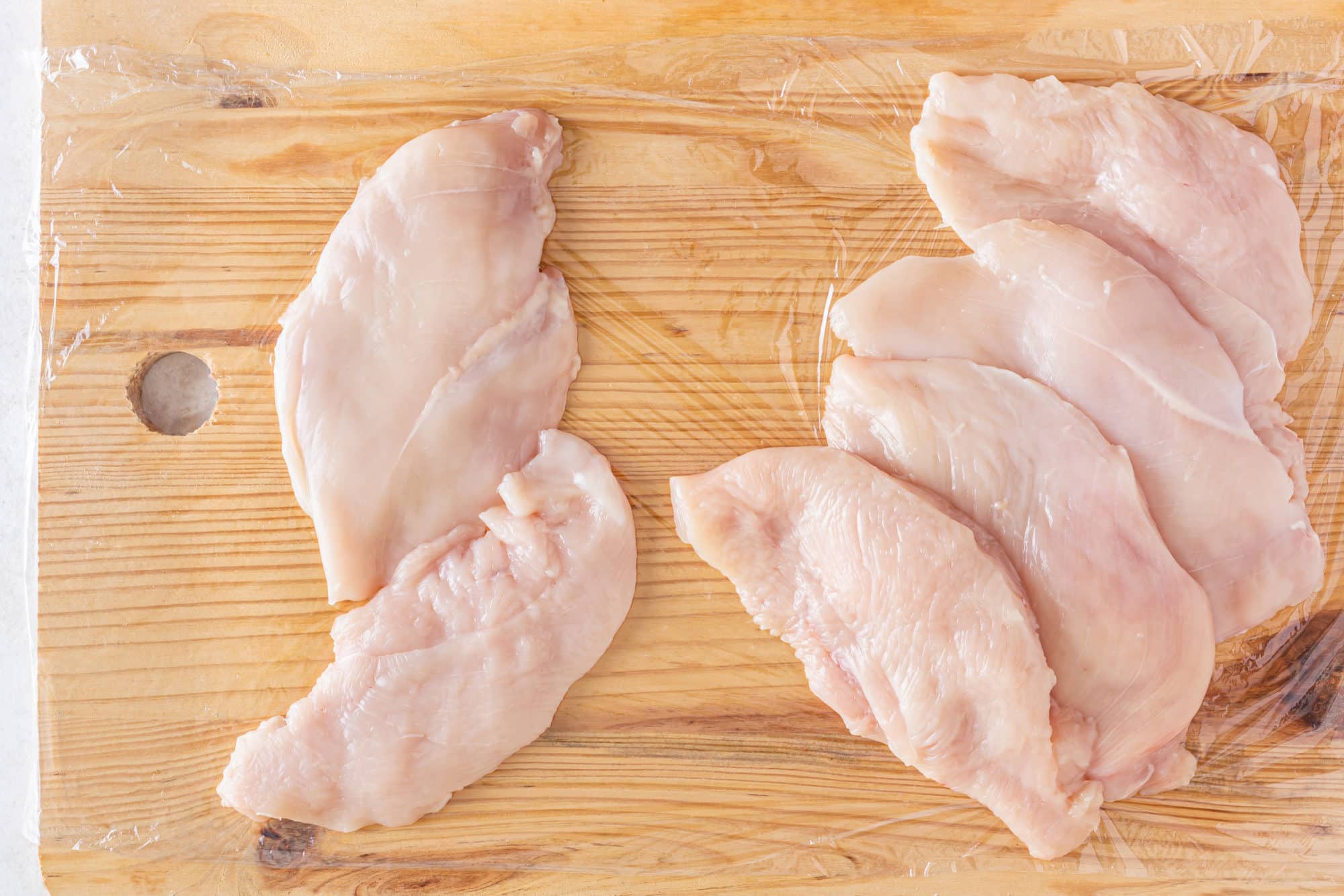 chicken breasts on a wooden board that is also covered in plastic wrap.