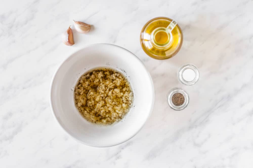 Garlic topping in a white bowl with garlic cloves, olive oil, salt, and pepper on the side.