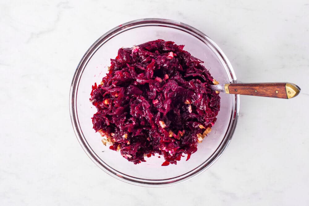 A glass bowl of shredded beets mixed with walnuts using a spoon.