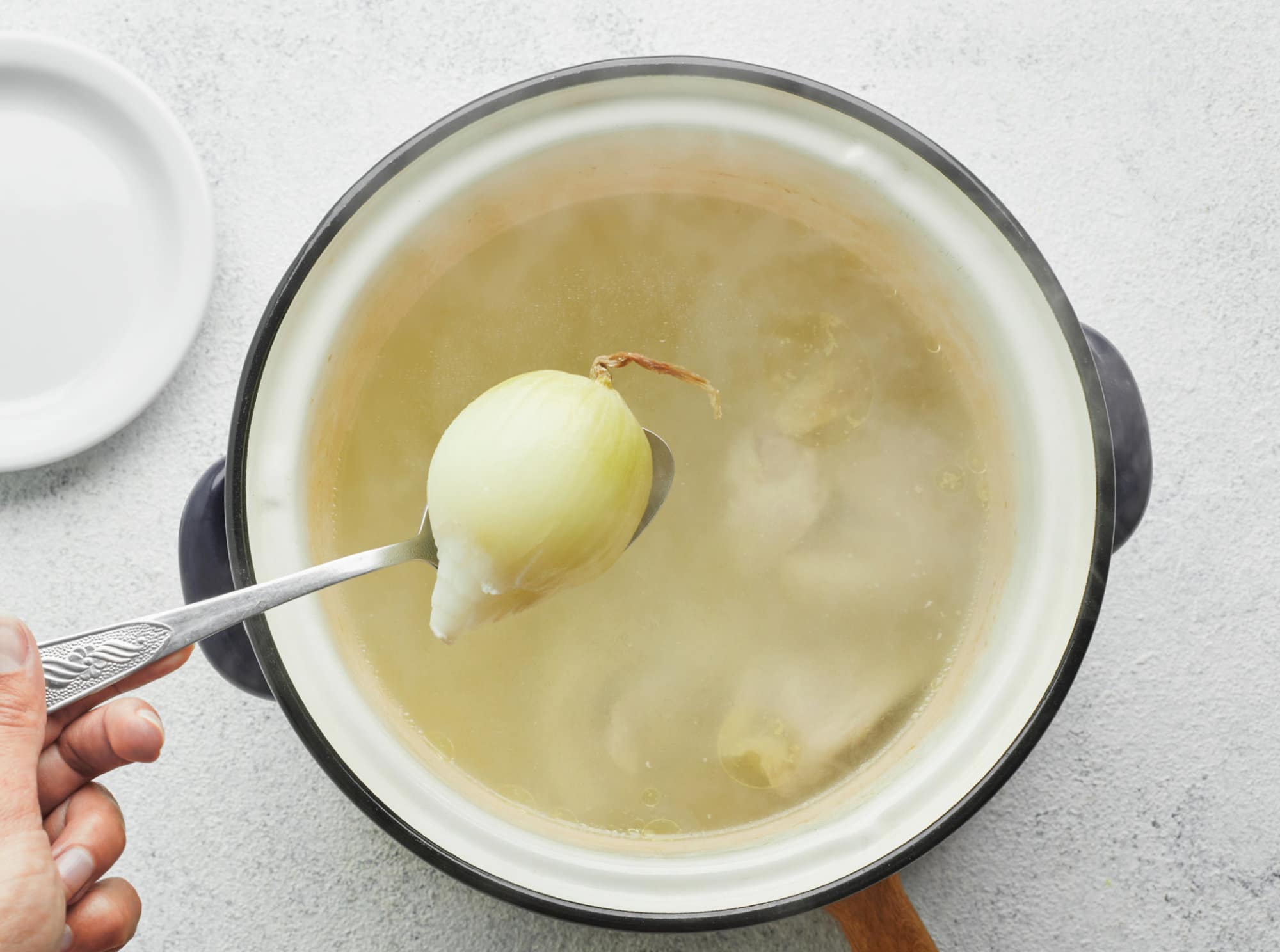 chicken-cooking-in-a-soup-pot-on-a-wooden-board-with-onion-being-added-in-with-a-spoon-with-a-white-plate-on-the-side