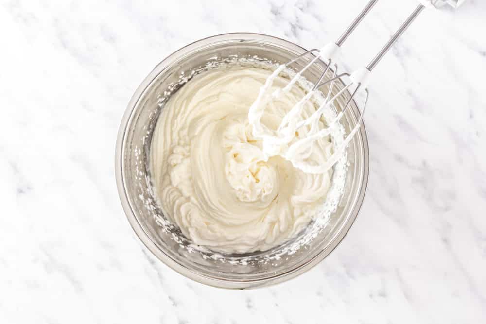 A silver bowl with whipped cream and silver hand mixer.