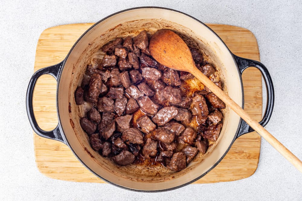 A wooden board with a pot on top of it with a wooden spoon and cooked meat in it.