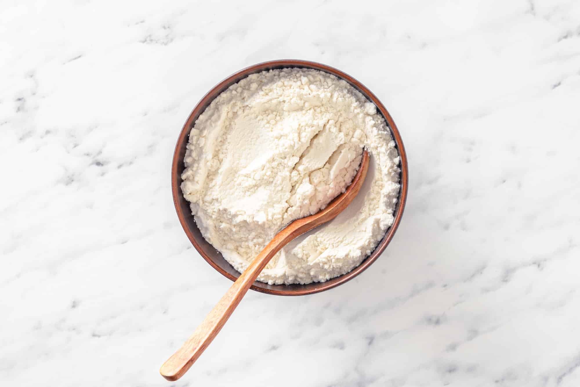 flour and other dry ingredients in a wooden bowl with a wooden spoon in the bowl.