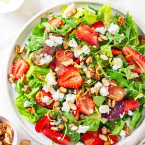 Summer berry salad on a white plate with a bowl of lemon zest and a bowl of almonds on the side.