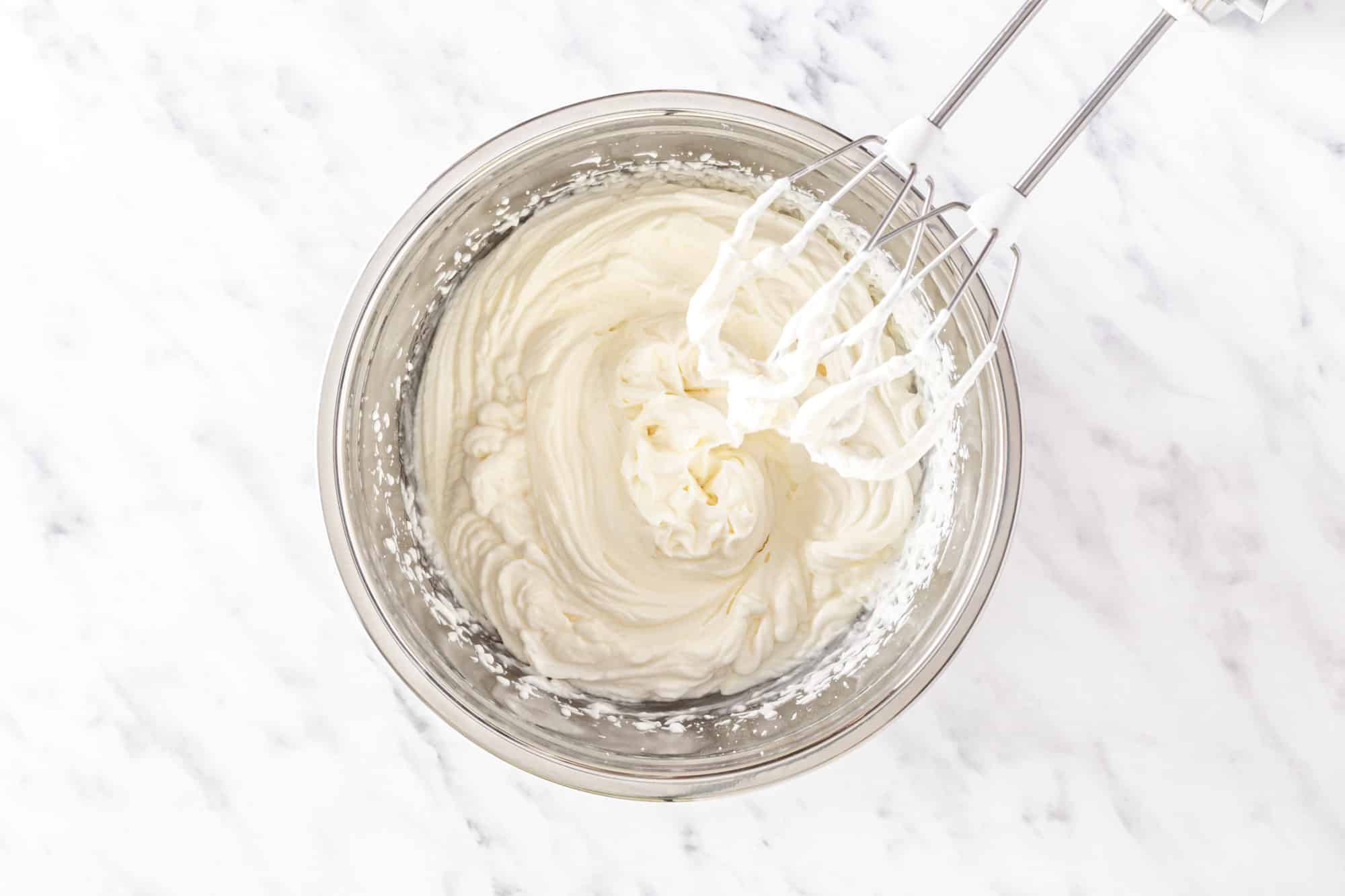 A silver bowl with whipped cream and silver hand mixer.