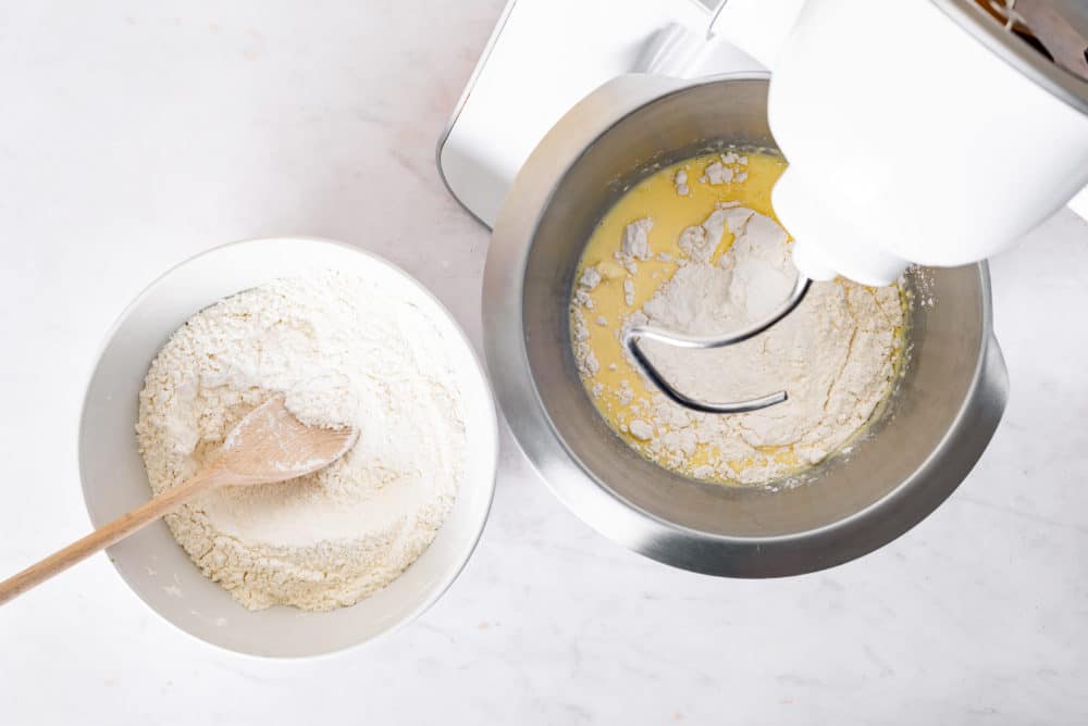 A bowl of flour and a wooden spoon and a mixer mixing dough for piroshki.