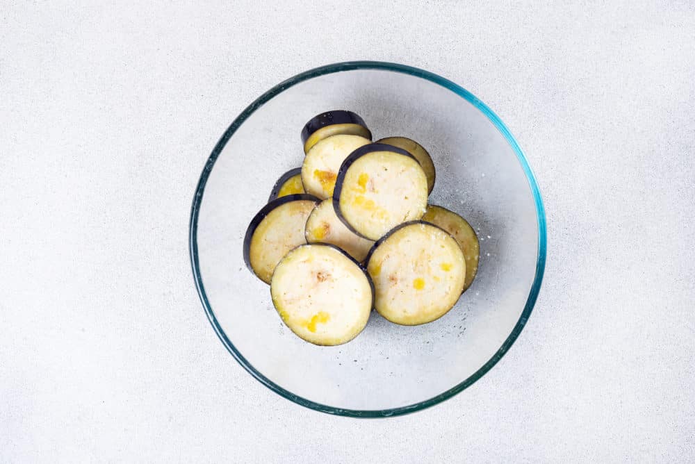 eggplant slices with olive oil salt and pepper mixed together in a glass bowl.