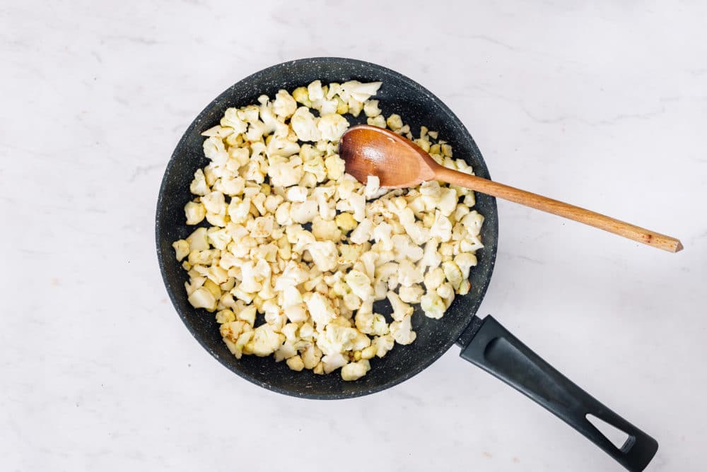 Cauliflower sauteing in a skillet with a wooden spoon.