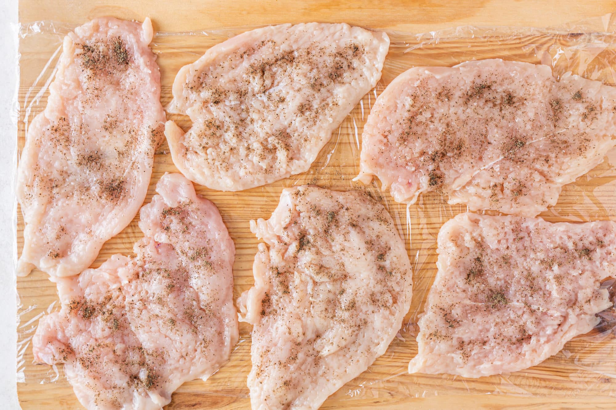 a wooden board covered in plastic wrap flattened chicken breasts and seasoning on top.