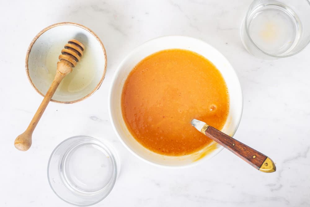 peach puree in a white bowl with a spoon in it and a honey dipper on the side and empty ingredient bowls.