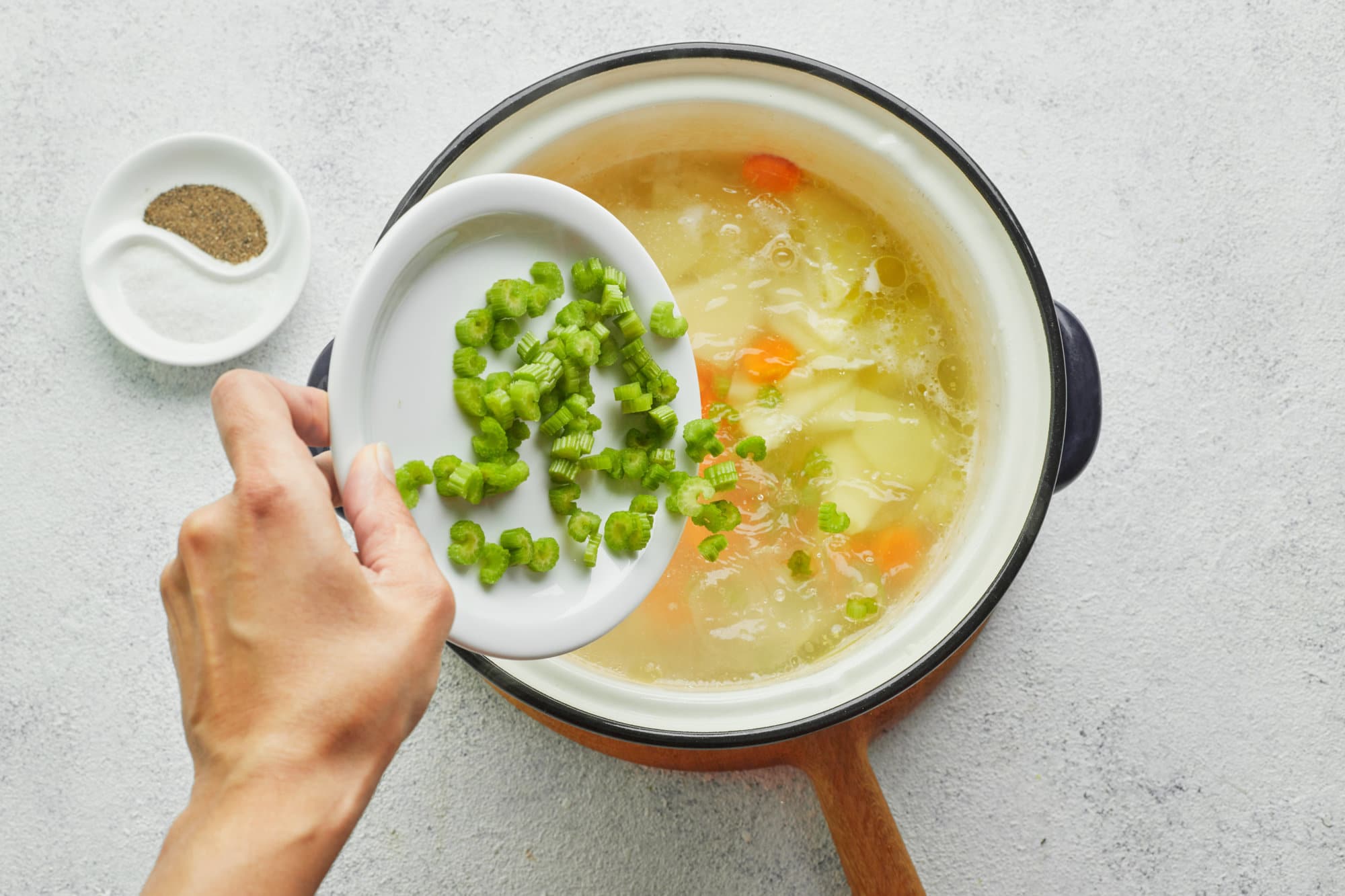 chicken-noodle-soup-in-a-soup-pot-on-a-wooden-board-with-green-onion-being-added-inside-with-a-bowl-of-salt-and-pepper-on-the-side