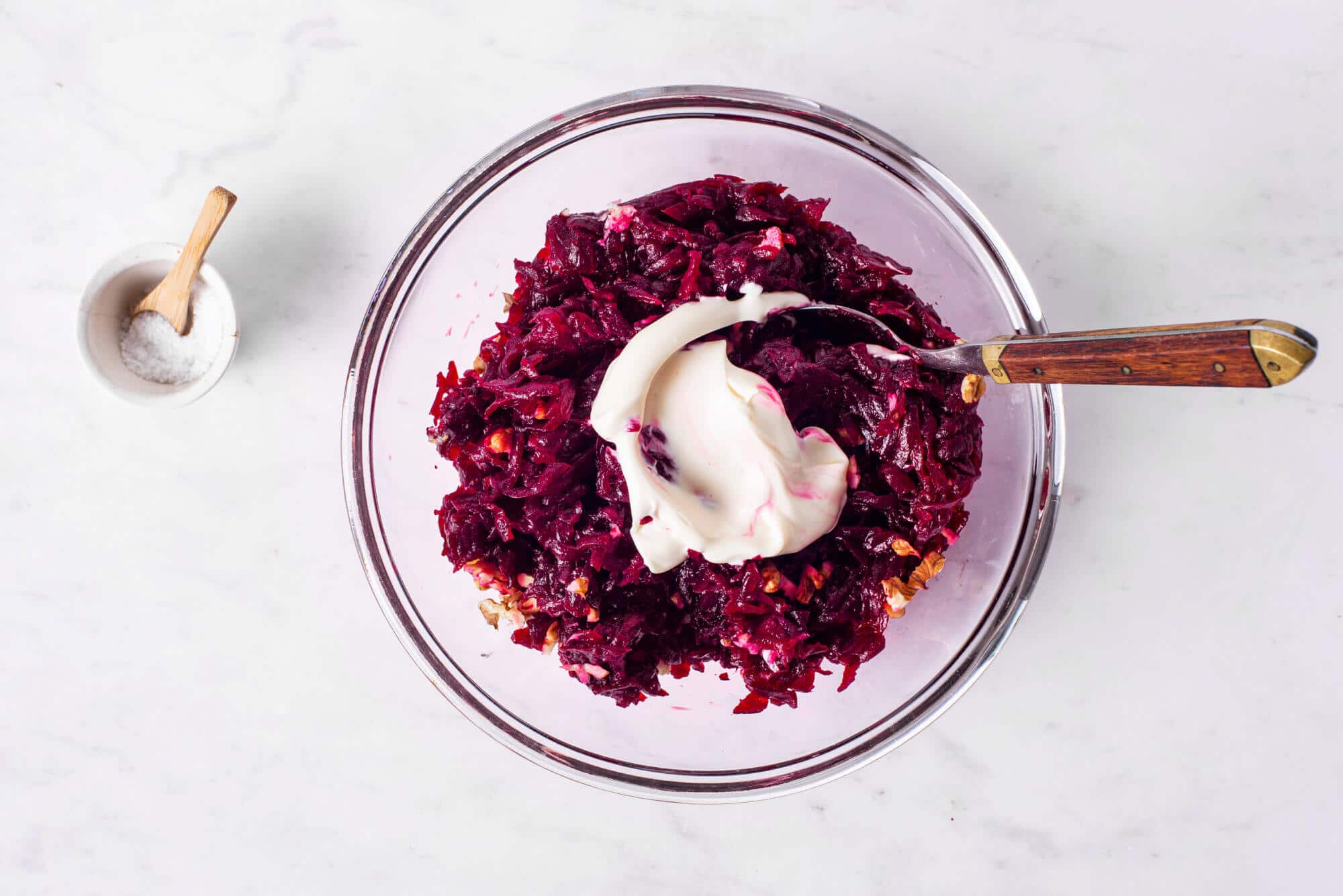 Mixing in mayonnaise into beet salad with a spoon, with salt on the side.