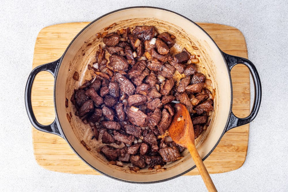 A wooden spoon in a pot with meat cooking in it on a wooden board.