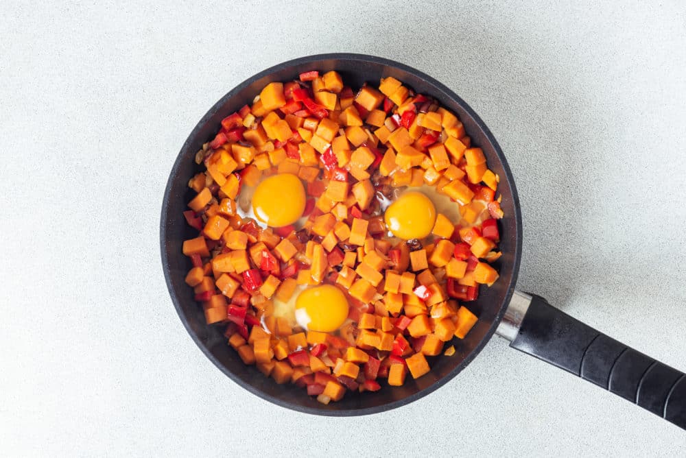 Sweet potatoes and red bell peppers with cracked raw eggs in a black skillet.