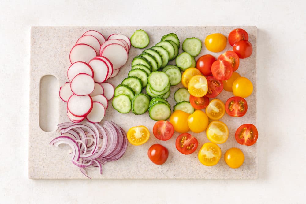 sliced radishes red onion tomato and cucumber on a cutting board.