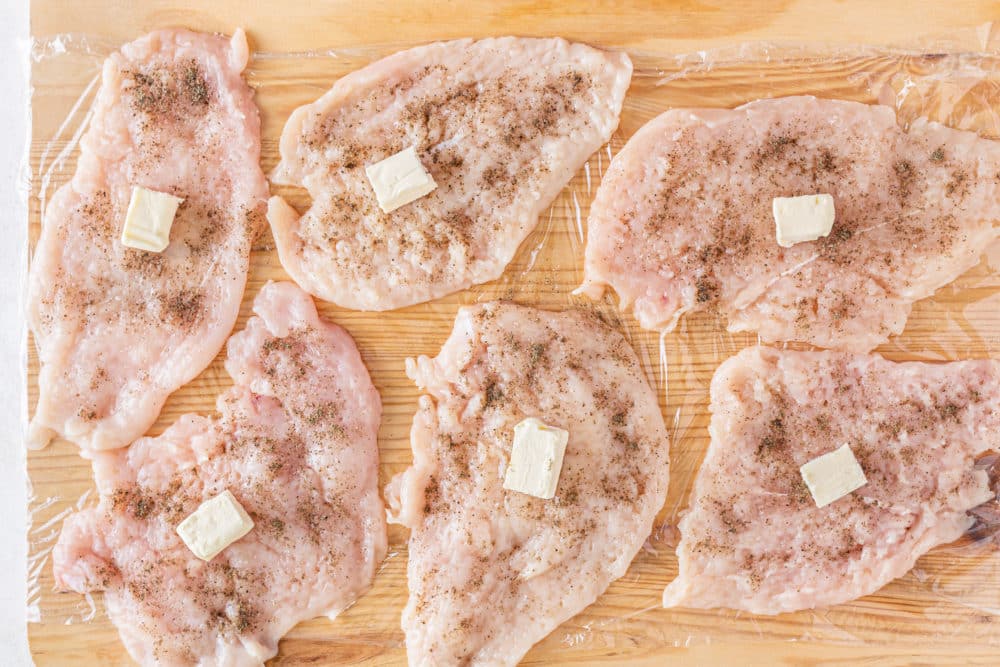 a wooden cutting board with plastic wrap over it with chicken breasts seasoning and butter on top.