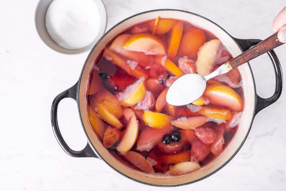 fruits and water in a pot adding sugar with a spoon and more sugar in a bowl on the side.