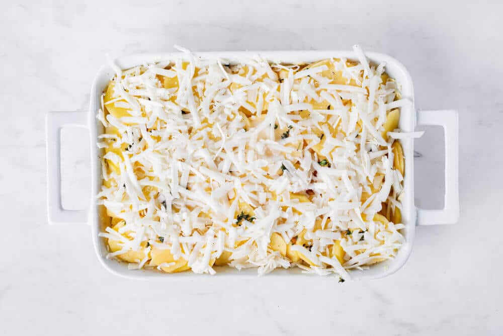 sliced potatoes in a white baking dish with seasoning and shredded cheese on top ready to bake.