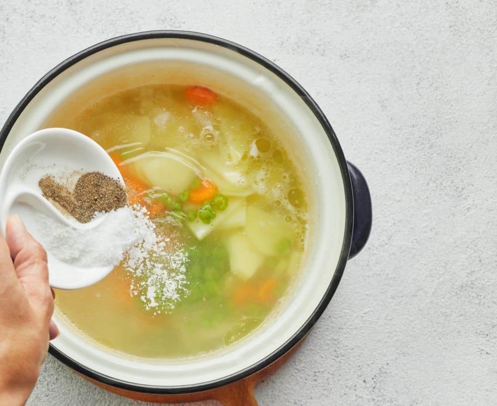 chicken-noodle-soup-in-a-soup-pot-on-a-wooden-board-with-salt-and-pepper-being-added-in-from-a-white-bowl