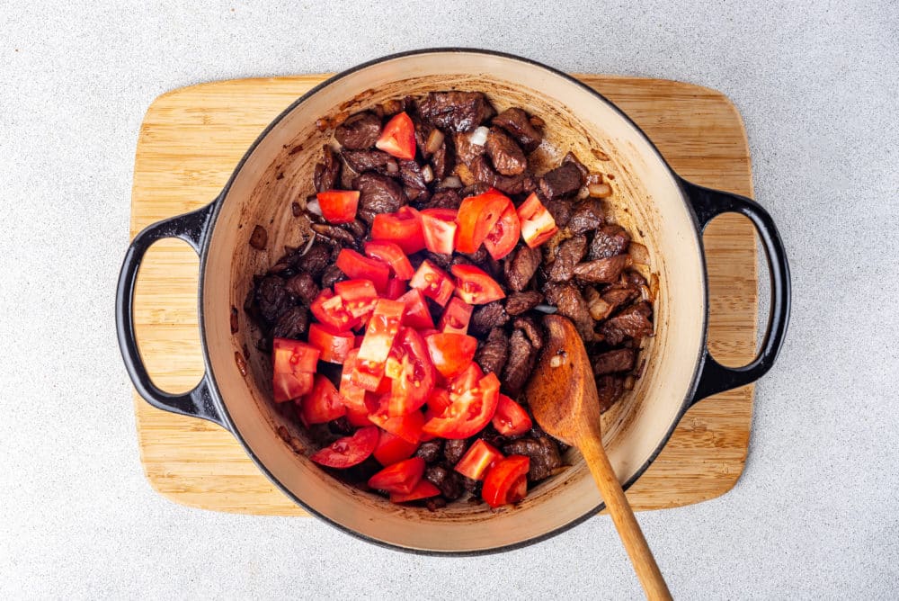 Beef and tomatoes in a large pot with a wooden spoon in the pot on a cutting board.