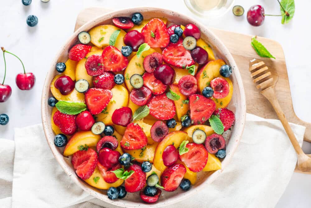 a summer fruit salad in a large bowl on a wooden board with a honey mixer on the side.