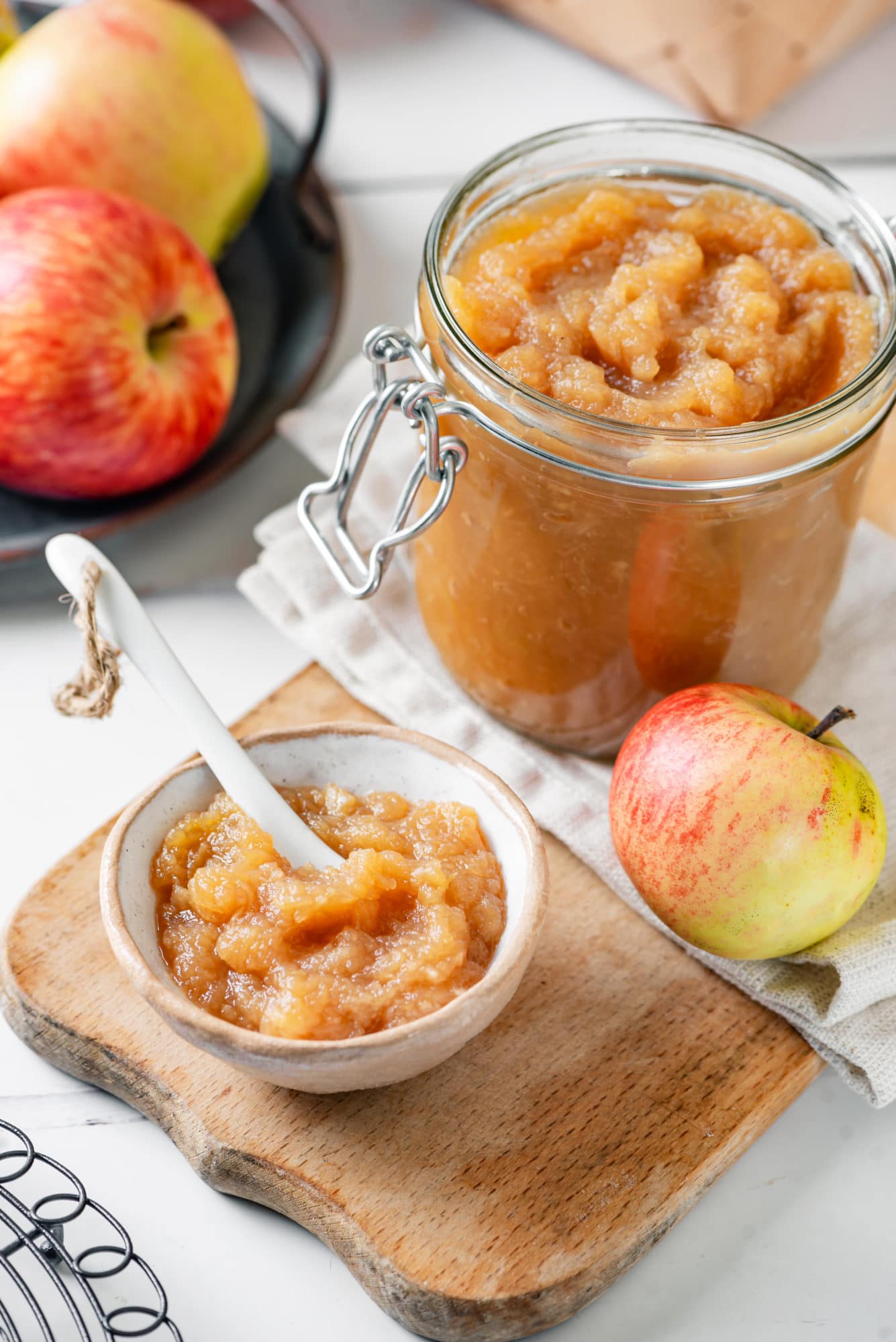 A glass jar and a white bowl of apple jam with a spoon in the bowl and apples all around.