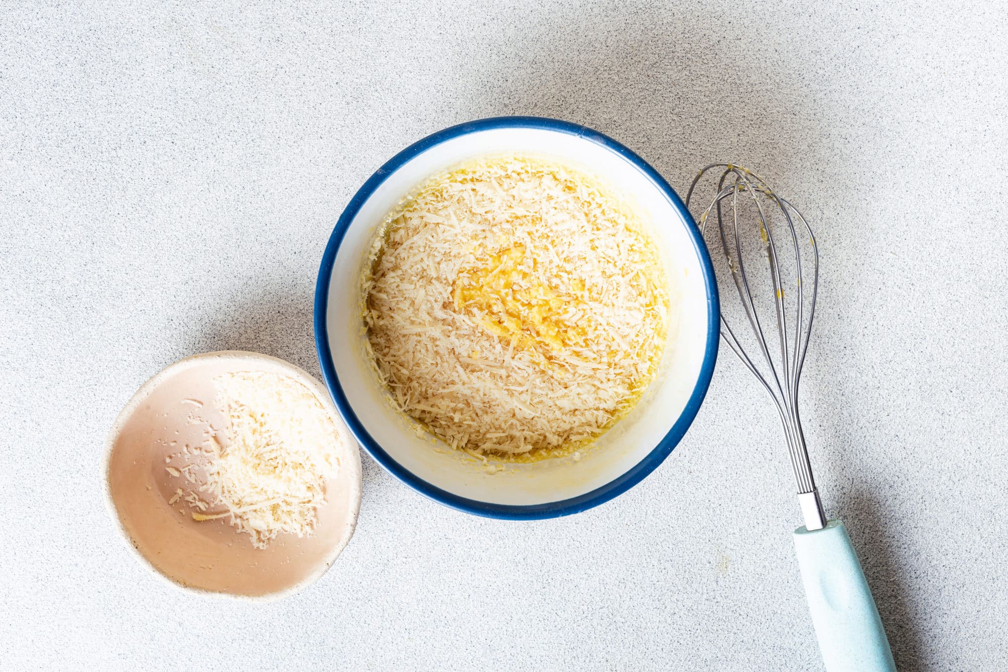 Whisked eggs in a white bowl with a whisk on the side and parmesan on top.