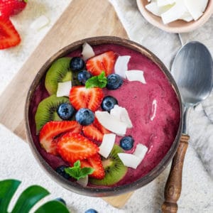 a purple acai smoothie bowl in a wooden bowl with a spoon on the side with fruit toppings on the smoothie bowl.