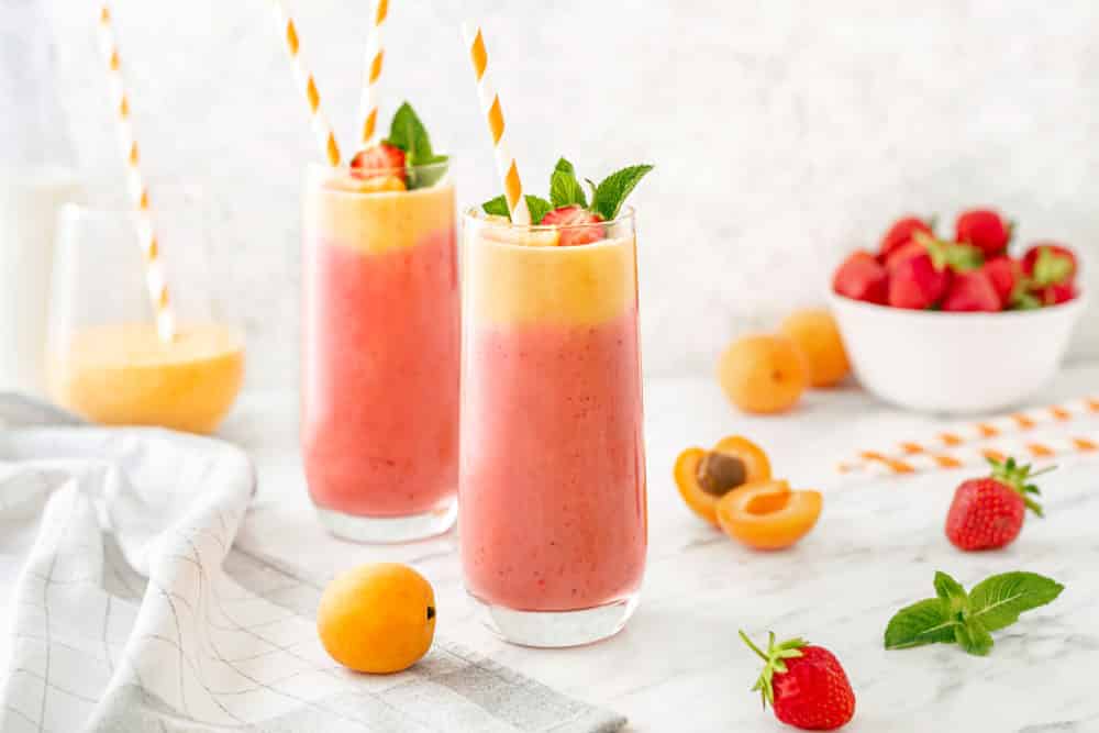 Two clear glasses filled with apricot and strawberry smoothie with striped straws topped with mint and fruit.