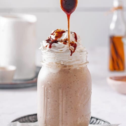 blended caramel smoothie in a mason jar with whipped cream on top and a spoon drizzling caramel.