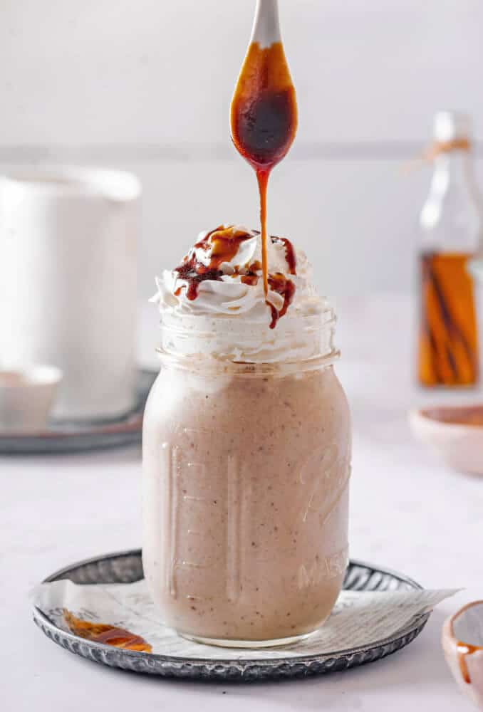 blended caramel smoothie in a mason jar with whipped cream on top and a spoon drizzling caramel.