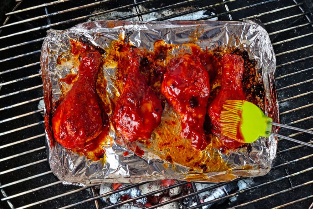 Chicken drumsticks on foil on a grill with a baster.