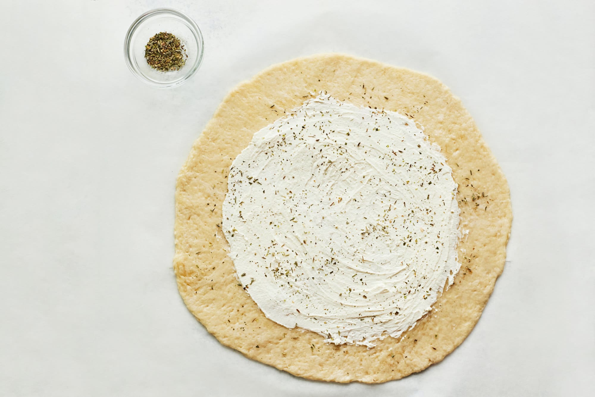 raw dough with cream cheese and italian seasoning with a bowl of seasoning on the side.