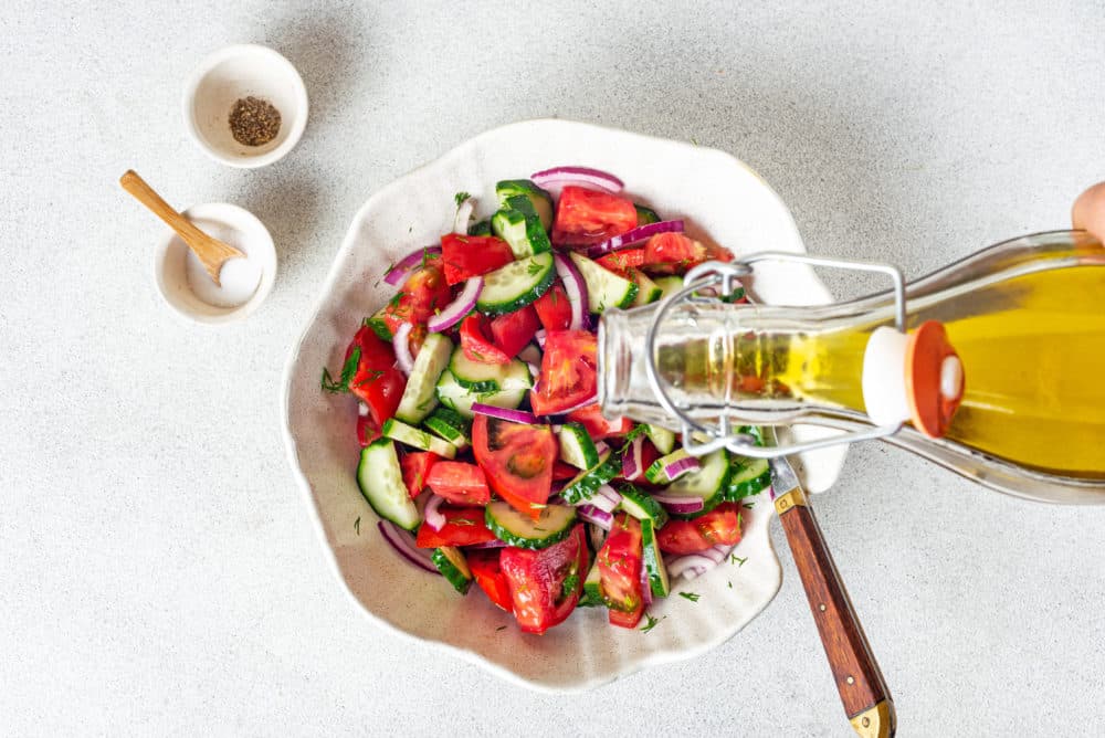 summer-salad-in-a-white-bowl-with-a-spoon-and-olive-oil-being-poured-into-the-bowl-and-salt-and-pepper-bowls-on-the-side