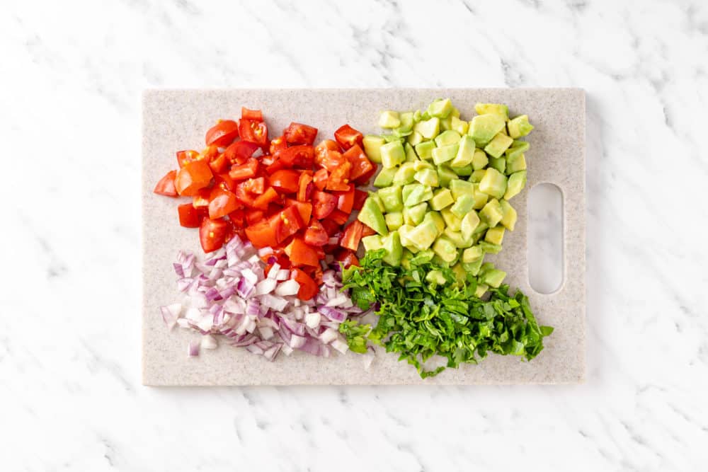 a cutting board with chopped tomatoes red onion avocado and parsley.